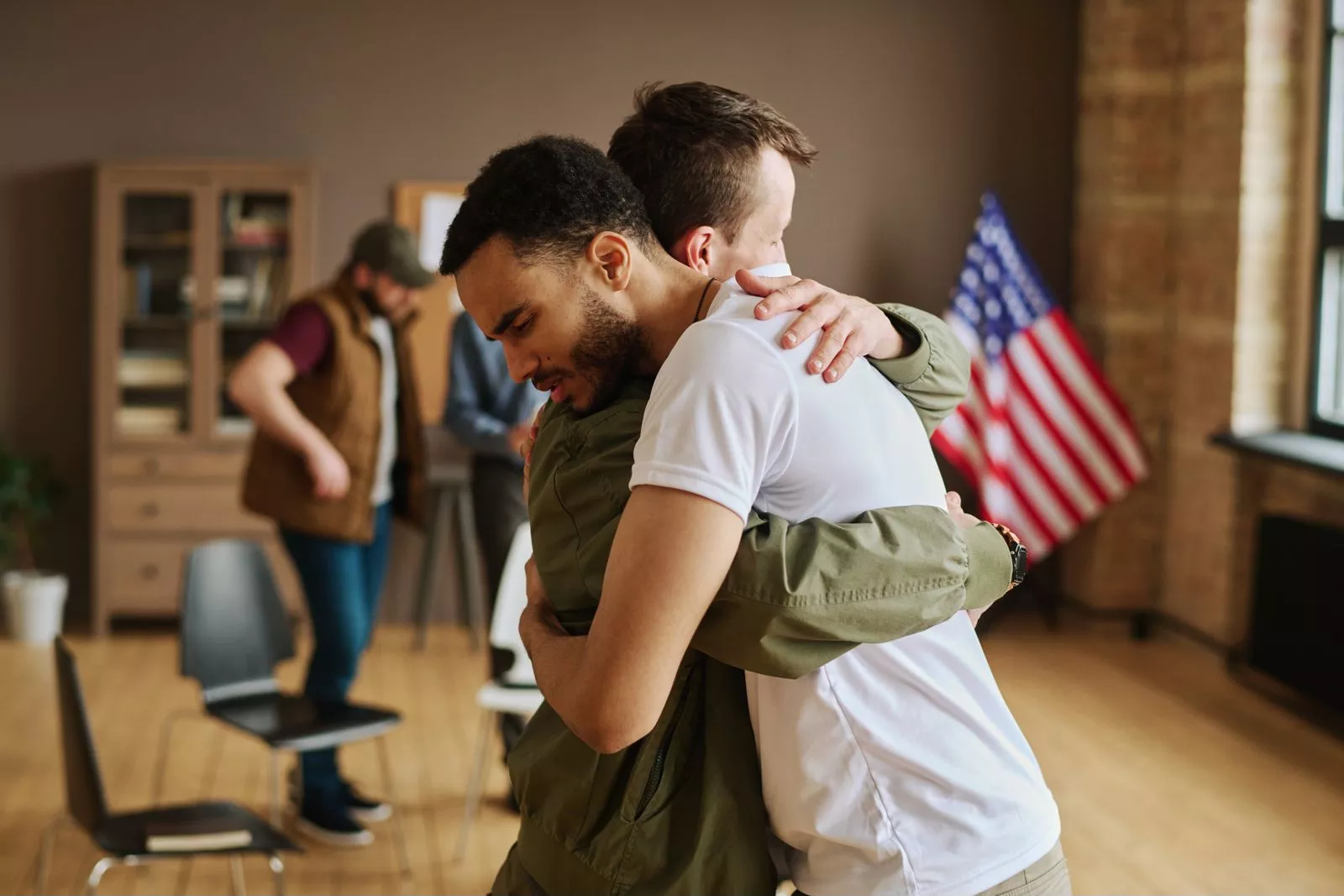 Homens se abraçam, um de jaqueta verde e outro de camiseta branca, em gesto de apoio. Bandeira dos EUA no fundo.