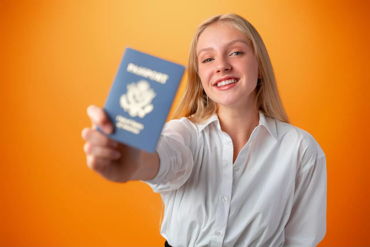 Mulher loira sorridente de camisa branca estende passaporte azul. Fundo laranja.