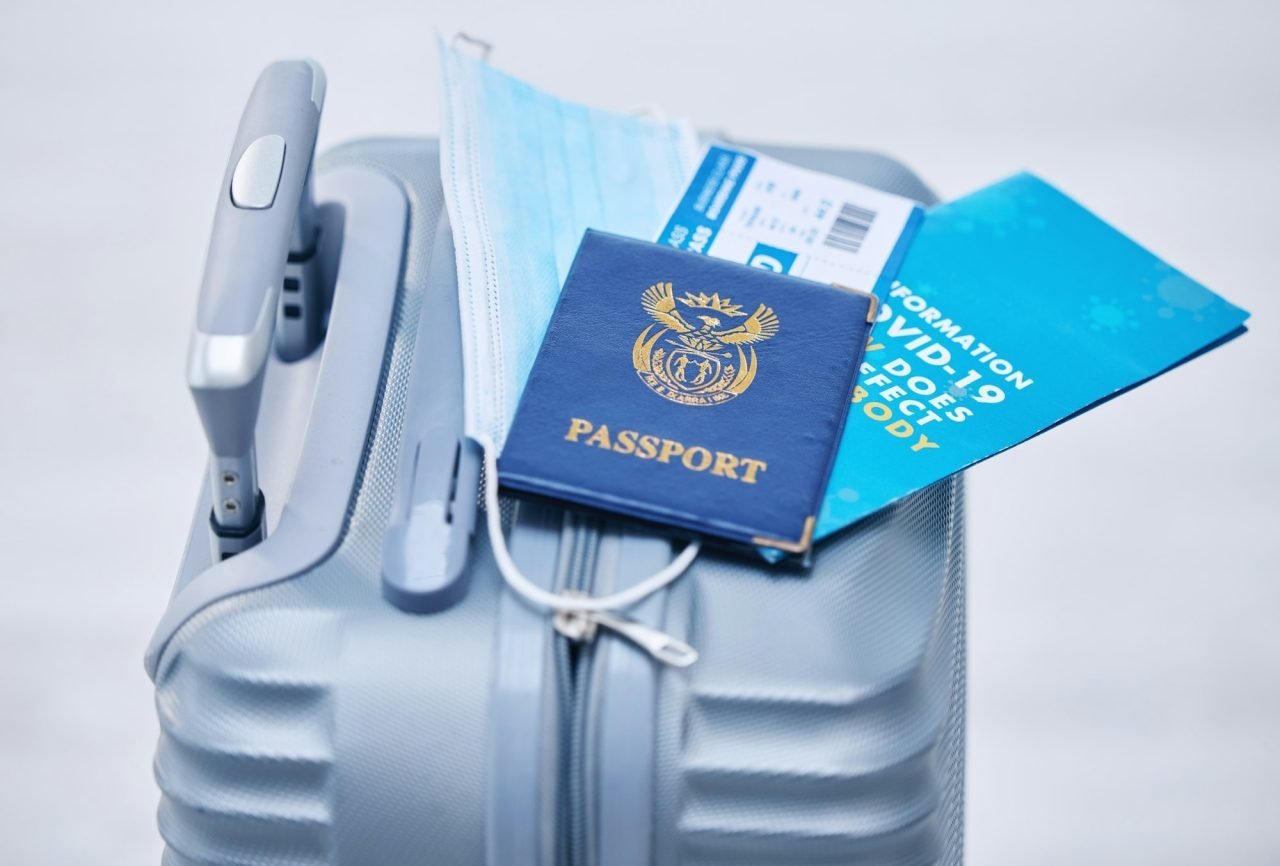 Mala de viagem cinza com passaporte azul, máscara facial, bilhete de avião e folheto sobre COVID-19.