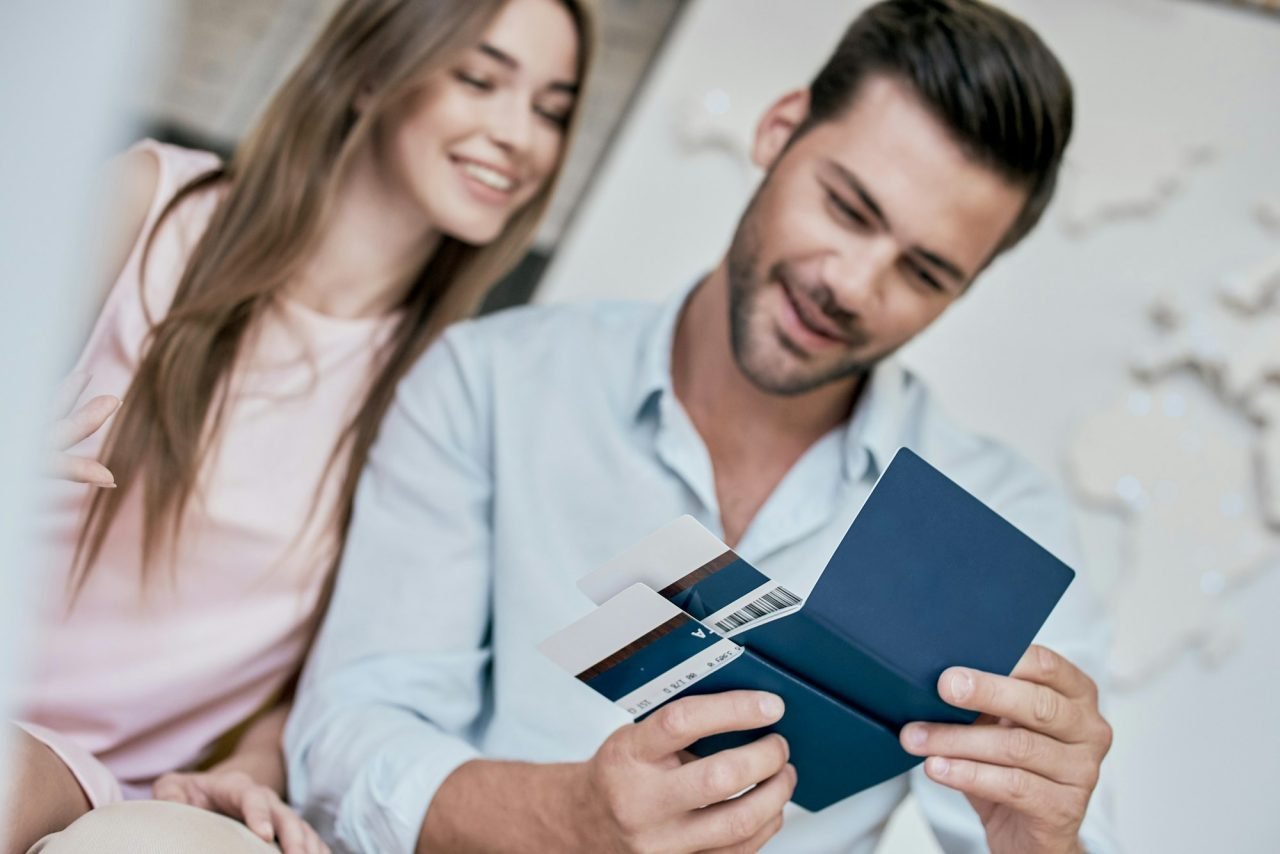 Casal feliz planeja viagem, segurando passaporte azul e passagens aéreas. Homem de camisa clara, mulher de blusa rosa.